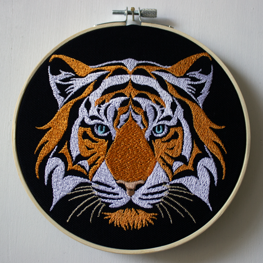 Embroidery of a tiger's face in an embroidery hoop on a plain background.